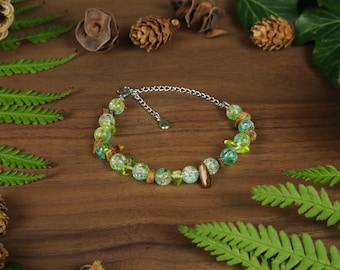 Könnte beinhalten: Ein Armband mit grünen und braunen Perlen und einem silbernen Kettenverschluss. Das Armband wird auf einer Holzoberfläche präsentiert, umgeben von grünen Farnblättern, Tannenzapfen und anderen natürlichen Elementen.