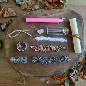 May include: A collection of spiritual items arranged on a wooden surface. Includes a pink candle, small glass bottles, a scroll, dried herbs, a crystal, and a small tin. The items are surrounded by dried leaves and flowers.