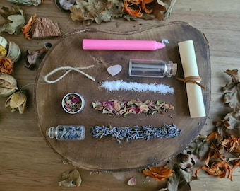 May include: A collection of spiritual items arranged on a wooden surface. Includes a pink candle, small glass bottles, a scroll, dried herbs, a crystal, and a small tin. The items are surrounded by dried leaves and flowers.