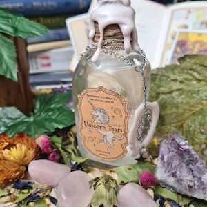 May include: A glass bottle with a white wax seal and a label that reads "Ravenwood Apothecary's one solution cure-all Unicorn Tears". The bottle is surrounded by pink and purple crystals, dried flowers, and green leaves.