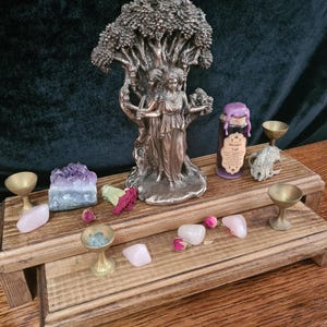 Peut inclure: Autel en bois à deux niveaux avec une statue en bronze de trois femmes debout sous un arbre, une petite bouteille étiquetée "L'Appel d'Hécate", et divers cristaux et autres objets décoratifs.
