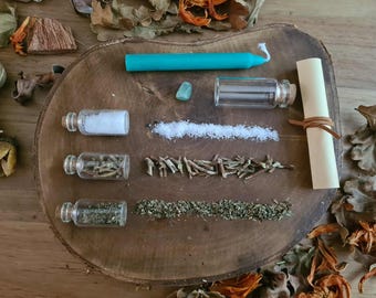 May include: A collection of items for spiritual practices, including a teal candle, small glass bottles with contents, a green stone, and a rolled parchment tied with leather. The items are arranged on a wooden surface with dried leaves and flowers.