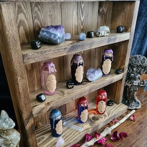 Pode incluir: Uma prateleira de madeira com duas prateleiras, exibindo várias garrafas de vidro com rótulos e selos de cera. As garrafas estão cheias de líquidos coloridos e são organizadas nas prateleiras com cristais e outros itens decorativos. A prateleira é feita de madeira rústica e tem um acabamento natural.