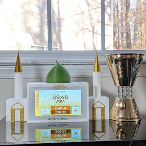 May include: A white Azan clock with a digital display showing the time and prayer information. The clock is flanked by decorative minaret-shaped structures with gold accents. A gold and silver incense burner is to the right.