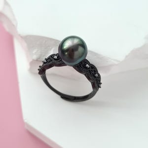 May include: A black ring with a large, dark gray pearl. The ring has an ornate, filigree design with small, clear accent stones. The ring is set against a white and pink background.