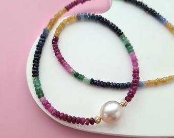 Könnte beinhalten: Eine mehrfarbige Edelsteinkette mit einem großen, runden, cremefarbenen Perlenanhänger. Die Kette besteht aus einer Vielzahl kleiner, farbiger Edelsteine, darunter Rot, Grün, Blau und Gelb. Die Kette wird auf einer weißen Oberfläche vor rosa Hintergrund präsentiert.