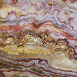 Puede incluir: Primer plano de una piedra pulida con un patrón en capas. La piedra presenta bandas de color rojo, amarillo, blanco y gris, creando un diseño natural y arremolinado. La superficie tiene un acabado liso y brillante, que resalta los intrincados detalles de la piedra.