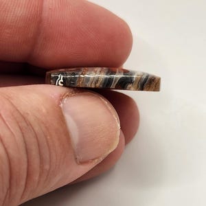 May include: A small, polished stone with a rectangular shape and rounded edges. The stone features a natural pattern of brown, black, and white bands. The stone is held between two fingers, showcasing its size and detail.