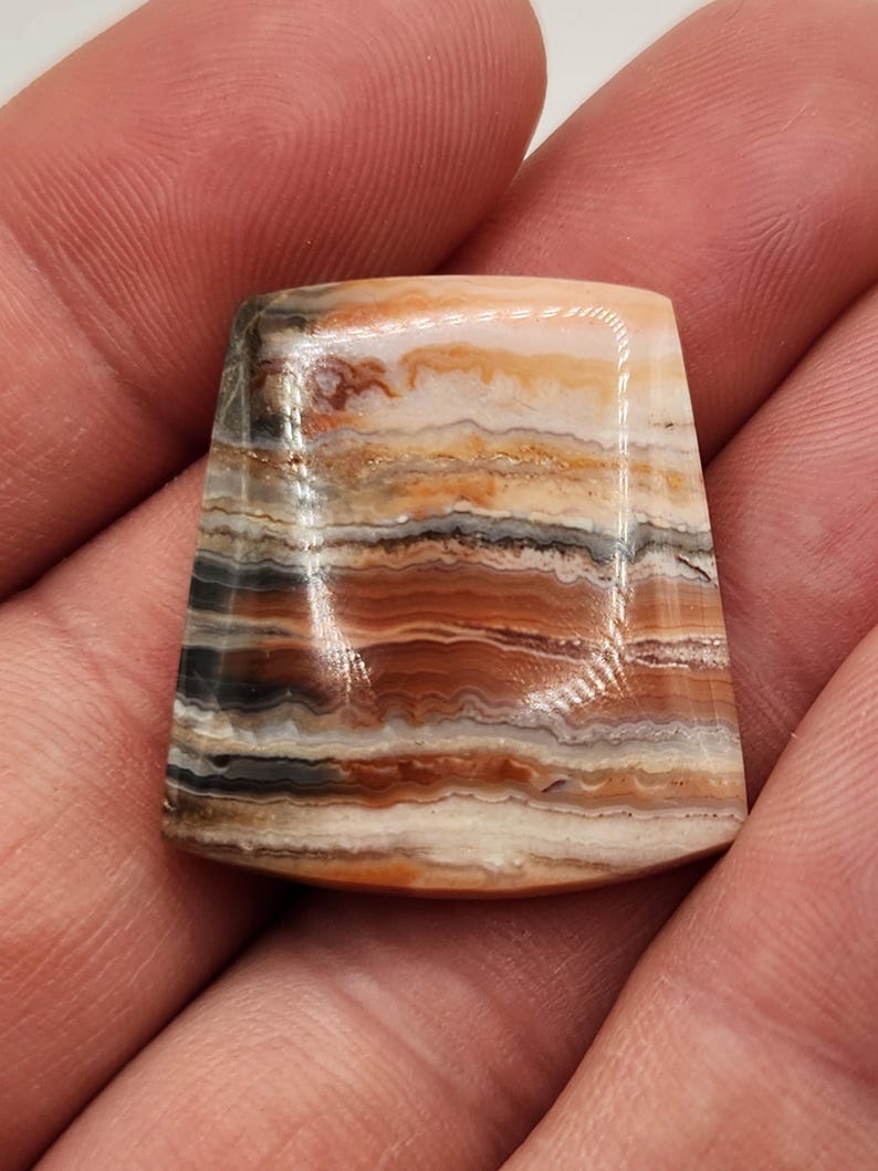 May include: A polished, trapezoid-shaped gemstone with a layered pattern of tan, brown, gray, and black bands. The stone has a smooth, glossy surface and is held in a hand, showcasing its natural colors and unique design.