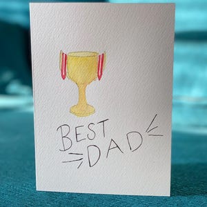 May include: A white card with a hand-drawn illustration of a golden trophy with red accents. The text "BEST DAD" is written below the trophy.