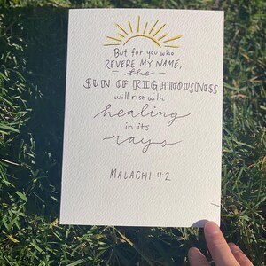 May include: A white card with handwritten text and a gold sun illustration. The text reads "But for you who revere my name, the sun of righteousness will rise with healing in its rays. Malachi 4:2."