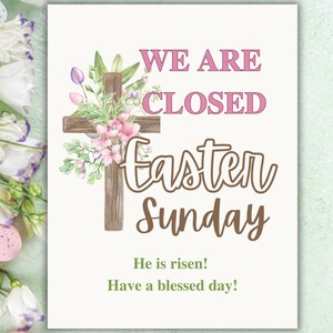 May include: A white sign with a watercolour illustration of a wooden cross with pink flowers. The sign reads "We are closed Easter Sunday. He is risen! Have a blessed day!"