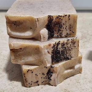 May include: Three bars of handmade soap with a light brown color and black poppy seeds. The soap is stacked on top of each other.