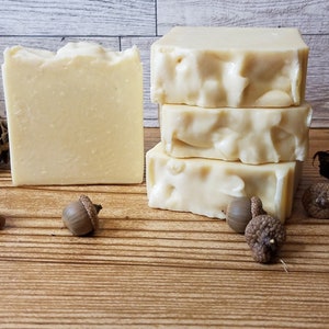 May include: Four bars of white soap stacked on a wooden surface. The soap is a light ivory color and has a slightly textured surface. There are acorns and pine cones scattered around the soap.