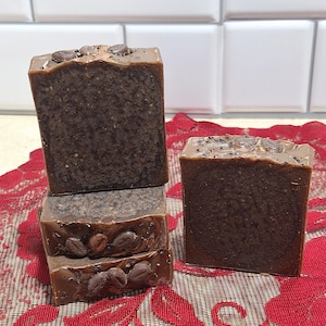 May include: Four bars of handmade coffee soap, stacked on top of each other. The soap is brown with coffee grounds and coffee beans embedded in it. The soap is sitting on a red lace doily.
