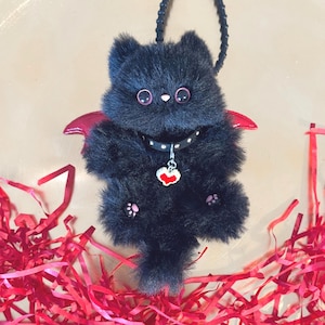 Könnte beinhalten: Ein schwarzes Plüschkätzchen mit roten Fledermausflügeln und rosa Pfoten. Die Katze trägt ein schwarzes Halsband mit einem Herzanhänger. Eine schwarze Schlaufe ist oben befestigt. Rote Papierschnipsel sind verstreut.