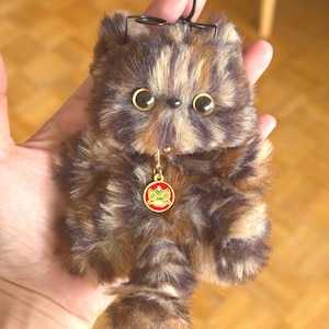 Peut inclure: Un porte-clés en forme de chat en peluche aux tons marron et beige, avec de grands yeux dorés et un petit pendentif rouge. Le porte-clés est doté d'un mousqueton et d'une armature métallique noirs. La fourrure du chat est douce et texturée.