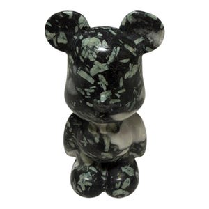 Può includere: Statuetta decorativa a forma di orso in posizione eretta. L'orso è realizzato in pietra maculata bianca e nera con accenti verdi. L'orso ha un corpo arrotondato, braccia piccole e grandi orecchie.