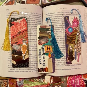 May include: Three colorful bookmarks with tassels. The first bookmark features a red and white illustration of a book with the text "Back to Hogwarts". The second bookmark features a blue and white illustration of a stack of books with the text "When in Doubt Go to the Library". The third bookmark features a blue and white illustration of a pair of glasses and a stack of books.