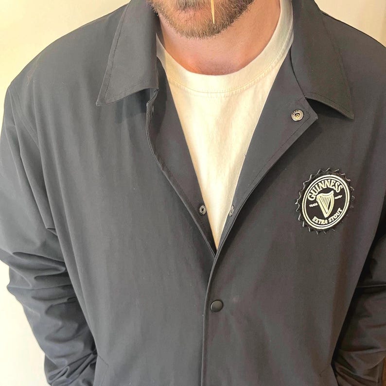 May include: A black jacket with a snap closure and a Guinness Extra Stout logo on the left side. The logo is a white circle with a harp inside and the words "Guinness Extra Stout" around it.