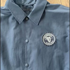 May include: A dark blue jacket with a black button closure and a white embroidered Guinness logo with the text "Guinness Extra Stout".