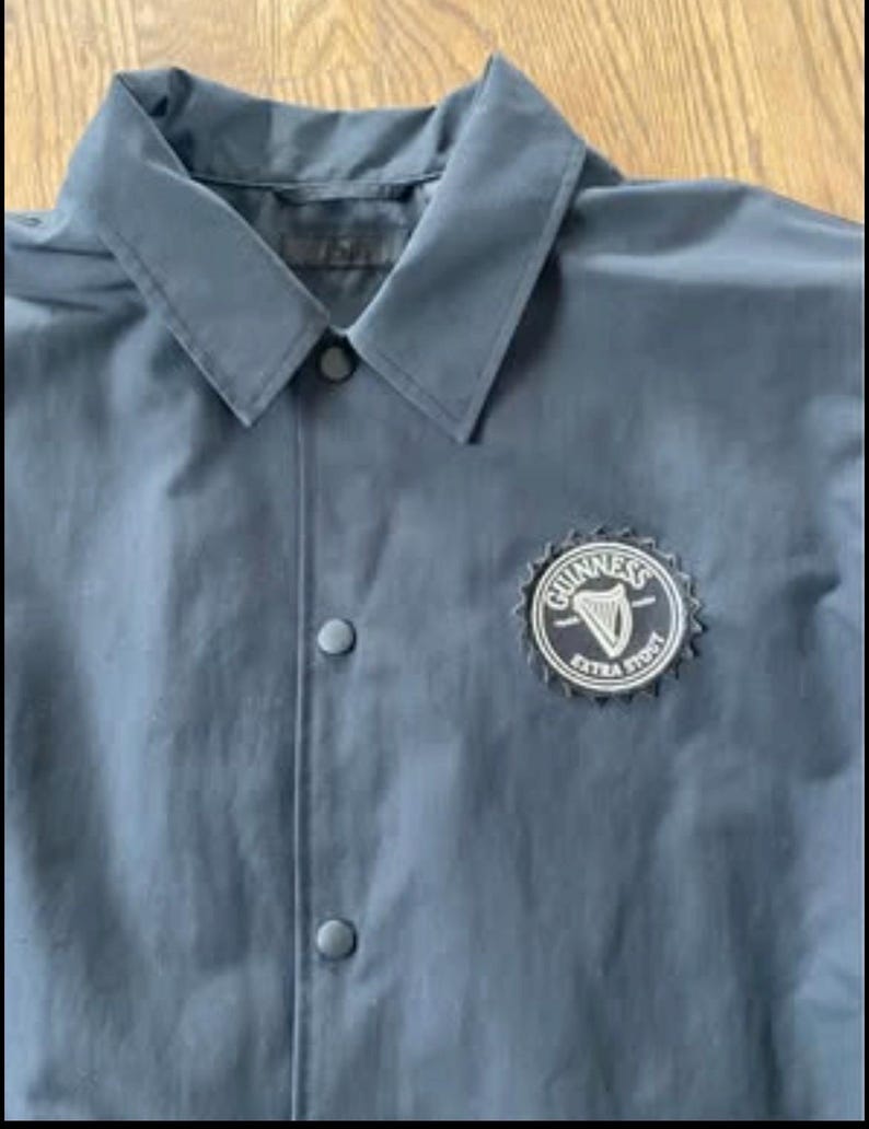 May include: A dark blue jacket with a black button closure and a white embroidered Guinness logo with the text "Guinness Extra Stout".