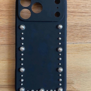 Puede incluir: Funda de teléfono negra con tachuelas plateadas dispuestas en los bordes. La funda tiene recortes para la cámara y otras funciones del teléfono. Las tachuelas son de diferentes tamaños, lo que añade un elemento decorativo.