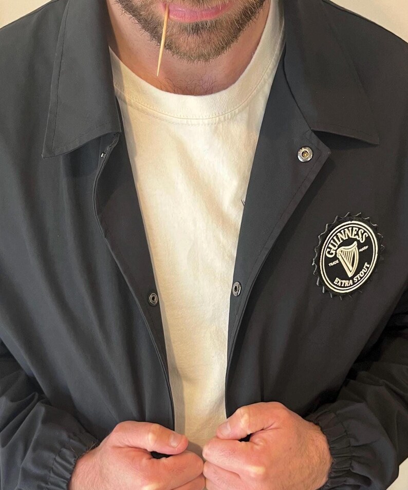 May include: A black jacket with a snap closure and a Guinness Extra Stout logo patch on the right side. The jacket has a white t-shirt underneath.