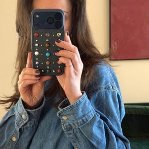 Puede incluir: Una funda de teléfono azul oscuro adornada con varias piedras preciosas y tachuelas de colores. La funda se sostiene frente a una persona que lleva una chaqueta vaquera azul. La funda tiene una cámara y un flash.