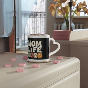 Puede incluir: Una taza de café negra con un asa en forma de corazón y las palabras "MOM LIFE" en blanco. La taza presenta iconos coloridos de un cochecito, una taza de café y una copa de vino. La taza está sobre un posavasos con caramelos rosas en forma de corazón.