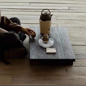 May include: A dark wooden tea table with a small teapot and teacup. A brown teapot sits atop a cream-colored warmer. A small, ornate teapot rests on a clear, cloud-shaped dish. A small, folded cloth is also on the table.