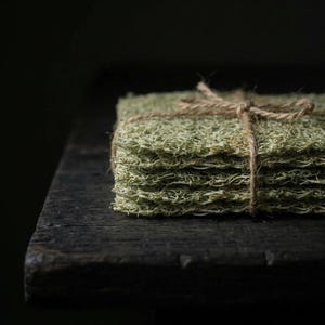 Op de afbeelding: Een stapel lichtgroene, getextureerde, vierkante items vastgebonden met touw. De items lijken gemaakt te zijn van een vezelachtig materiaal, mogelijk natuurlijk. Het touw is om de stapel gewikkeld en vastgeknoopt met een strik. De items liggen op een donker houten oppervlak.