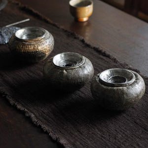 Puede incluir: Tres portavelas artesanales de cerámica con interior plateado metálico. Los portavelas son de color gris oscuro con superficies texturizadas y se encuentran sobre un camino de mesa tejido marrón oscuro. Una pequeña taza de té de color beige se encuentra en el fondo.