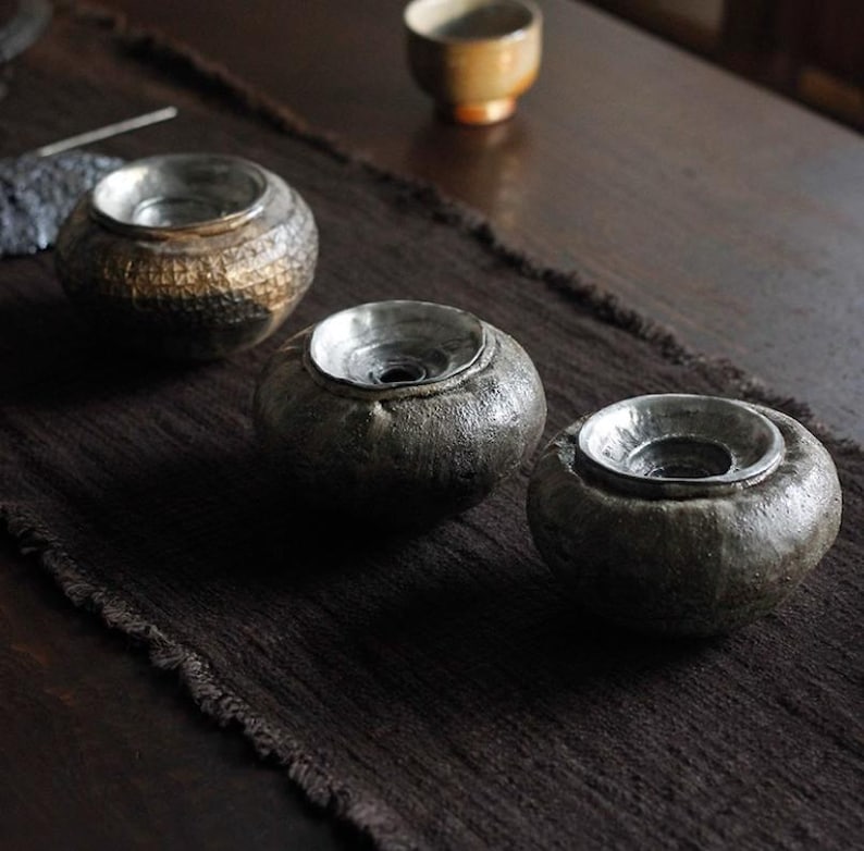 May include: Three handcrafted ceramic tea light holders with a metallic silver interior. The holders are dark gray with textured surfaces and sit on a dark brown woven runner. A small, tan teacup is in the background.