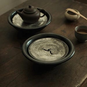 以下が含まれることがあります： ダークブラウンのセラミックティーポットと蓋が、シルバーのインサートが付いた黒いボウルに入っています。木のスプーンと小さなティーカップも木製の表面にあります。ティーセットは茶道用にデザインされています。