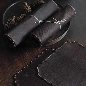 May include: Dark brown leather placemats and rolled napkins tied with twine. The napkins are rolled and tied with twine. The placemats have a decorative stitched edge. The items are on a dark wooden surface.