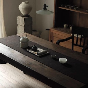 Puede incluir: Una mesa de madera oscura con un camino de mesa negro exhibe un juego de té, que incluye una tetera blanca, una tetera negra pequeña y un cuenco blanco. Un jarrón grande y texturizado se encuentra sobre un pedestal de piedra. También se ven un mueble y una silla de madera.