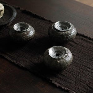 May include: Three small, dark brown ceramic tea cup rests with a silver-colored interior. The rests are arranged on a dark brown textile runner, with a wooden table in the background. The tea cup rests have a textured surface.
