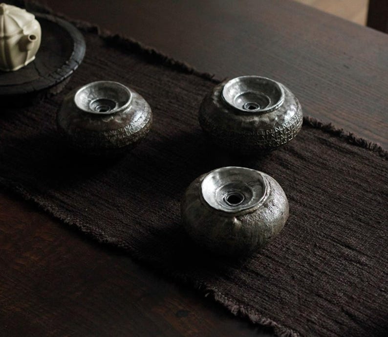 May include: Three small, dark brown ceramic tea cup rests with a silver-colored interior. The rests are arranged on a dark brown textile runner, with a wooden table in the background. The tea cup rests have a textured surface.