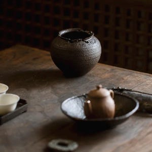 May include: A dark brown, textured ceramic tea set on a wooden table. The set includes a small teapot, a bowl, and a decorative jar with a ruffled rim. The background is a dark, patterned wall.