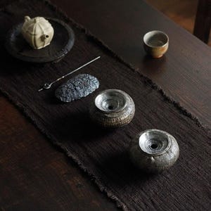 May include: A tea set arranged on a dark brown runner. The set includes a small, cream-colored teapot on a black plate, two ceramic vessels, a small cup, and a metal tool. The vessels have a textured, speckled appearance.