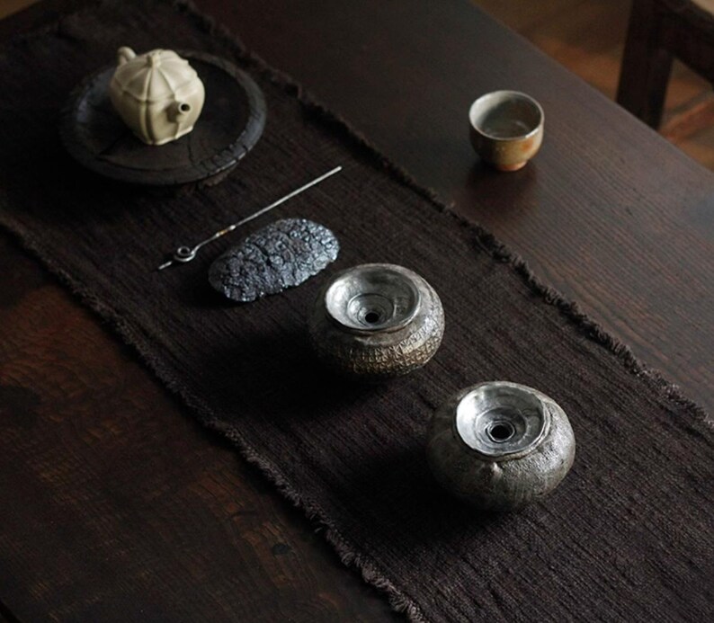 May include: A tea set arranged on a dark brown runner. The set includes a small, cream-colored teapot on a black plate, two ceramic vessels, a small cup, and a metal tool. The vessels have a textured, speckled appearance.