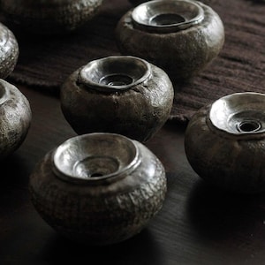 May include: A collection of small, dark gray ceramic bowls with a textured exterior and a smooth, silver-colored interior. The bowls have a rounded shape with a wide, shallow opening. The bowls are arranged on a dark brown surface.