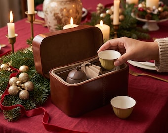 Puede incluir: Un estuche de transporte para juego de té de cuero marrón con tapa abatible, que contiene una pequeña tetera marrón oscuro y divisores de tela beige. Se incluyen dos tazas de té de cerámica clara con exterior acanalado.