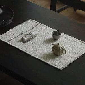 以下が含まれることがあります： 暗い木製のテーブルに置かれた穏やかなティーセット。小さな、明るい色の長方形のプレースマットには、小さなセラミックティーポット、カップ、装飾用の石が置かれています。暗いティーポットは背景の黒いトレイにあります。