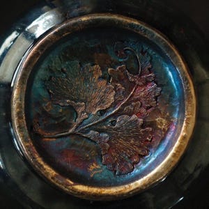 以下が含まれることがあります： 暗く虹色のボウルの内側のクローズアップ。イチョウの葉のデザインがエンボス加工されています。葉はブロンズと銅の色合いで、ティールと紫色の背景に配置されています。リムはメタリックゴールド仕上げです。