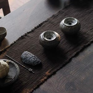 May include: A still-life arrangement featuring two textured, gray ceramic bowls with a central opening, set on a dark brown runner. A dark gray, stone-like object and a metal tool are also present, suggesting a tea ceremony setting.