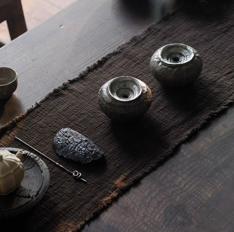 May include: A still-life arrangement featuring two textured, gray ceramic bowls with a central opening, set on a dark brown runner. A dark gray, stone-like object and a metal tool are also present, suggesting a tea ceremony setting.