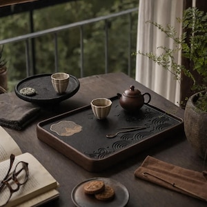 Könnte beinhalten: Ein hölzernes Teeservice mit einem dunkelbraunen Tablett, einer kleinen Teekanne und zwei Tassen. Das Tablett hat ein geschnitztes Wellenmuster und ein dekoratives Element. Das Set steht auf einem dunklen Holztisch mit einem Buch und einem kleinen Teller mit Keksen.