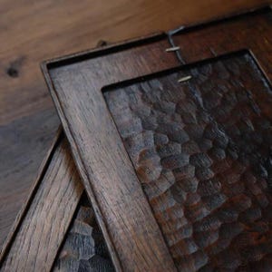 以下が含まれることがあります： 彫刻されたテクスチャのある表面を持つ、2つの濃い茶色の木製トレイ。トレイは長方形で縁が高く、手作りのようです。木目が見え、トレイはわずかに傾けて積み重ねられています。